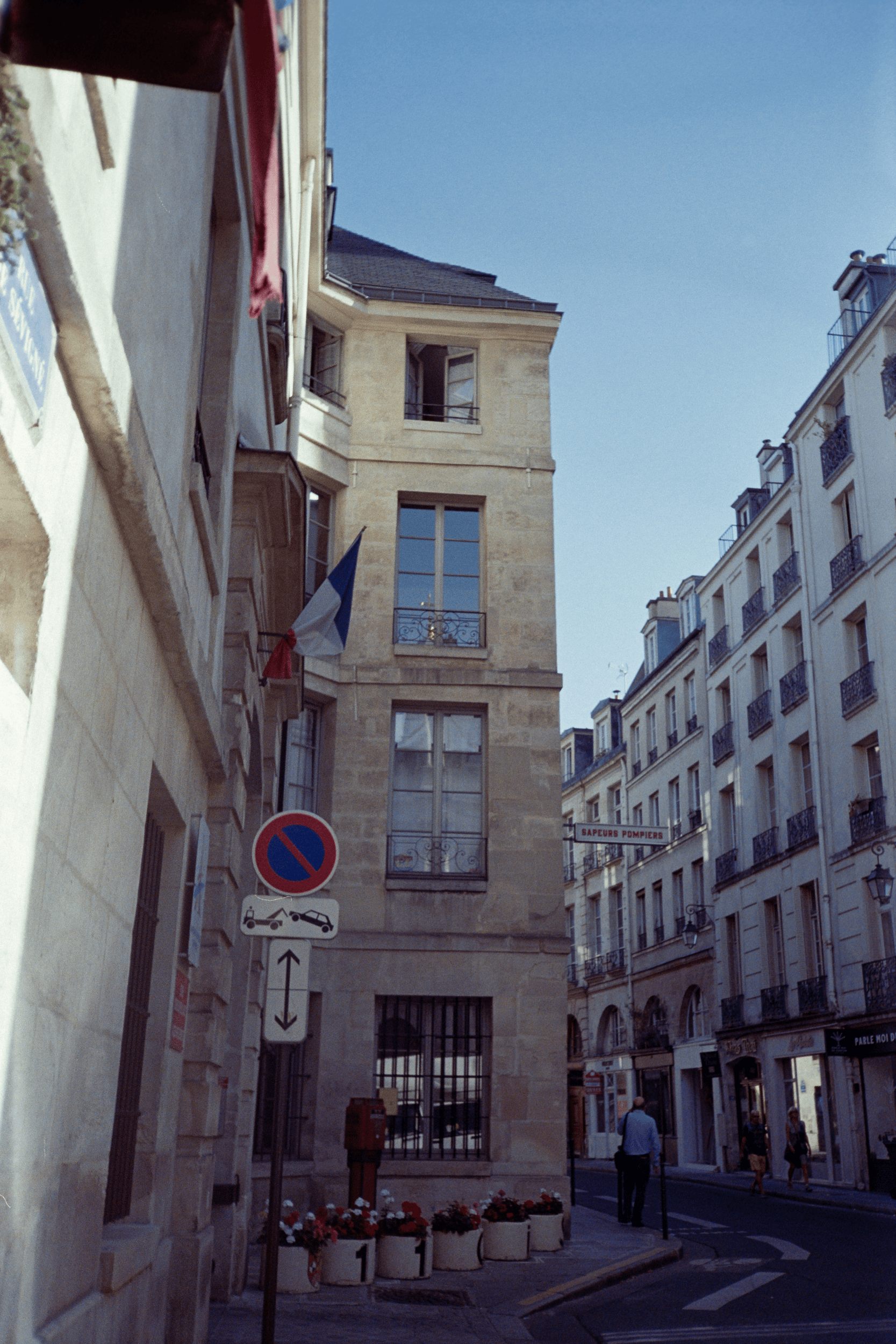 A film photo of a street view in Paris, focused on the edge of a building which the street curves around. The building is in partial shadow, a French flag hangs out of one of the windows.